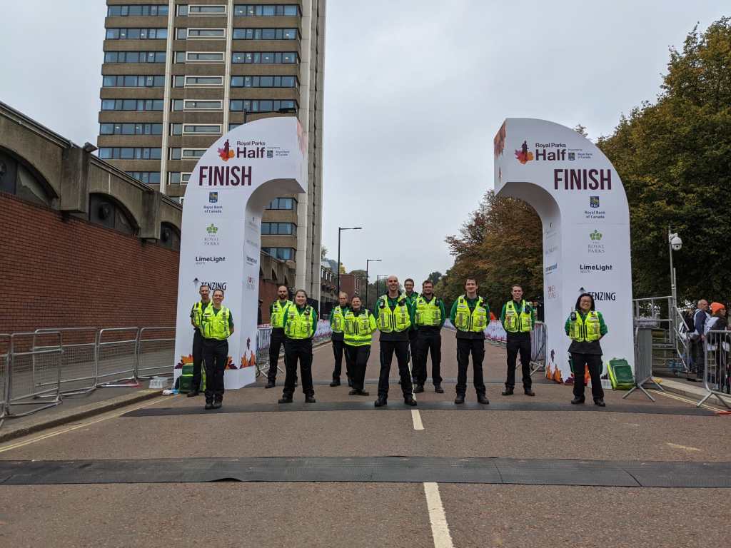 Twelve people in load-bearing vests standing formally in front of an arch marking the finish line of the Royal Parks Half Marathon